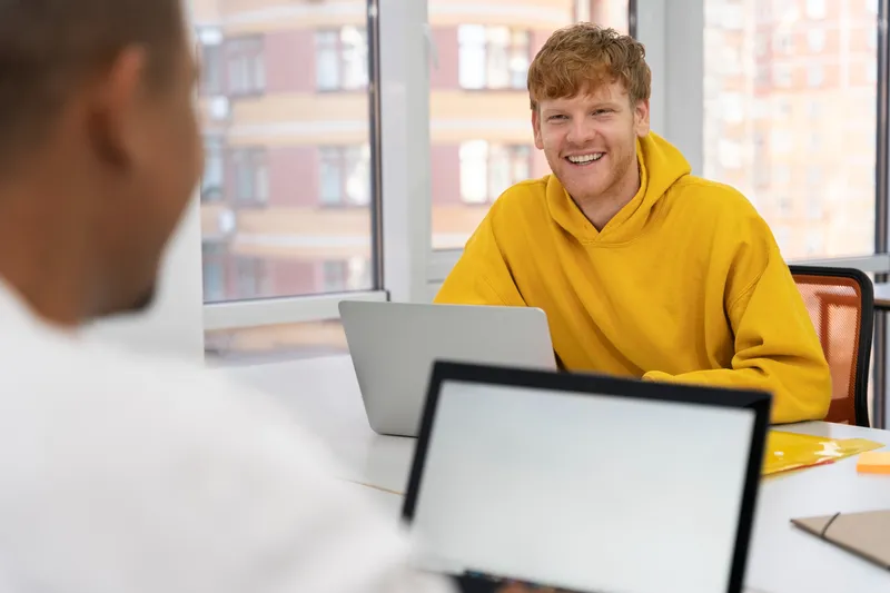 A colleague in a yellow sweater talks with someone across a desk by the window